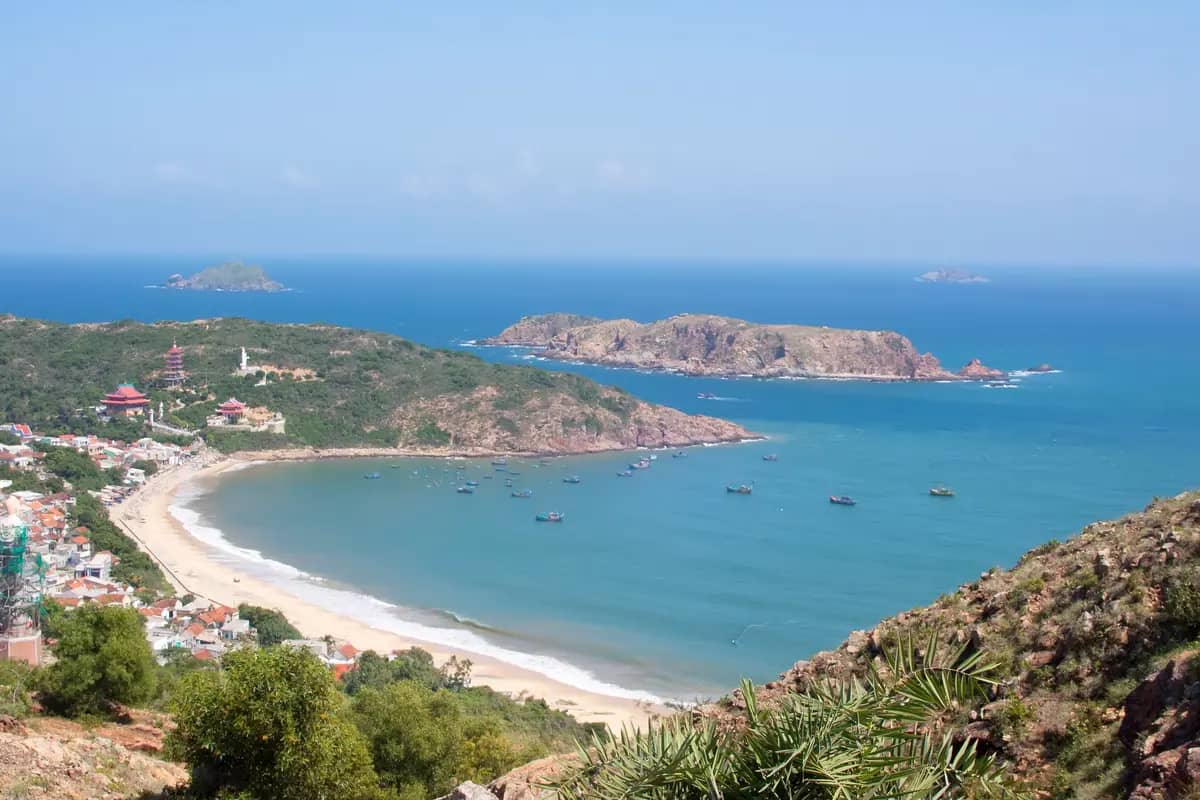 Ky Co bay near Quy Nhon seen from a hillside: a curved sandy beach, fishing boats on turquoise water and rocky islands