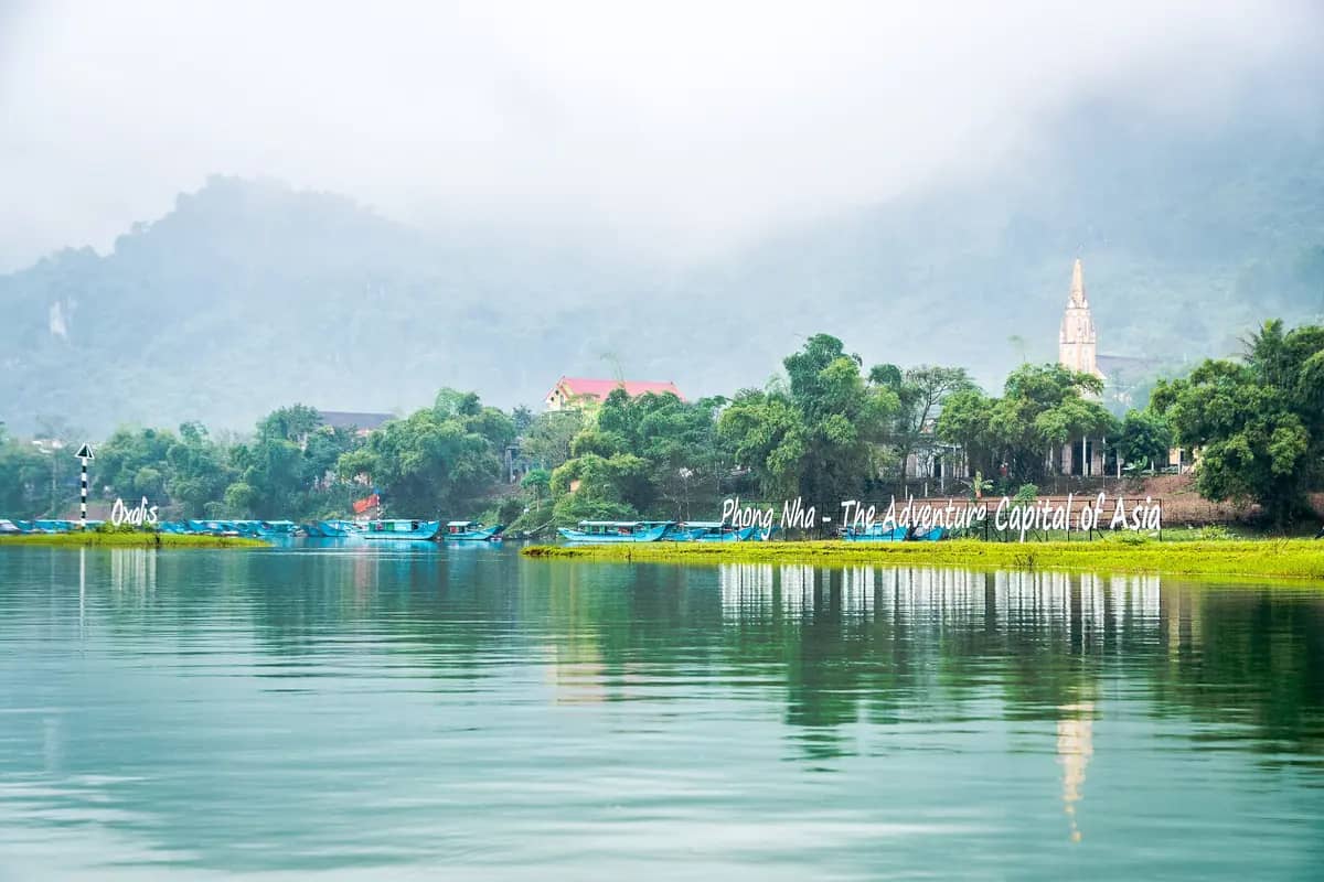 The Son River waterfront in Phong Nha town, tour boats moored below a church spire and misty hills