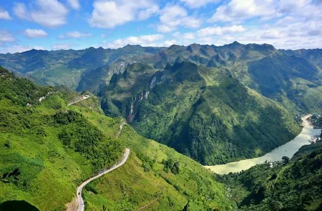 A narrow road winds along green Ma Pi Leng slopes above the jade Nho Que River, karst peaks under a blue sky