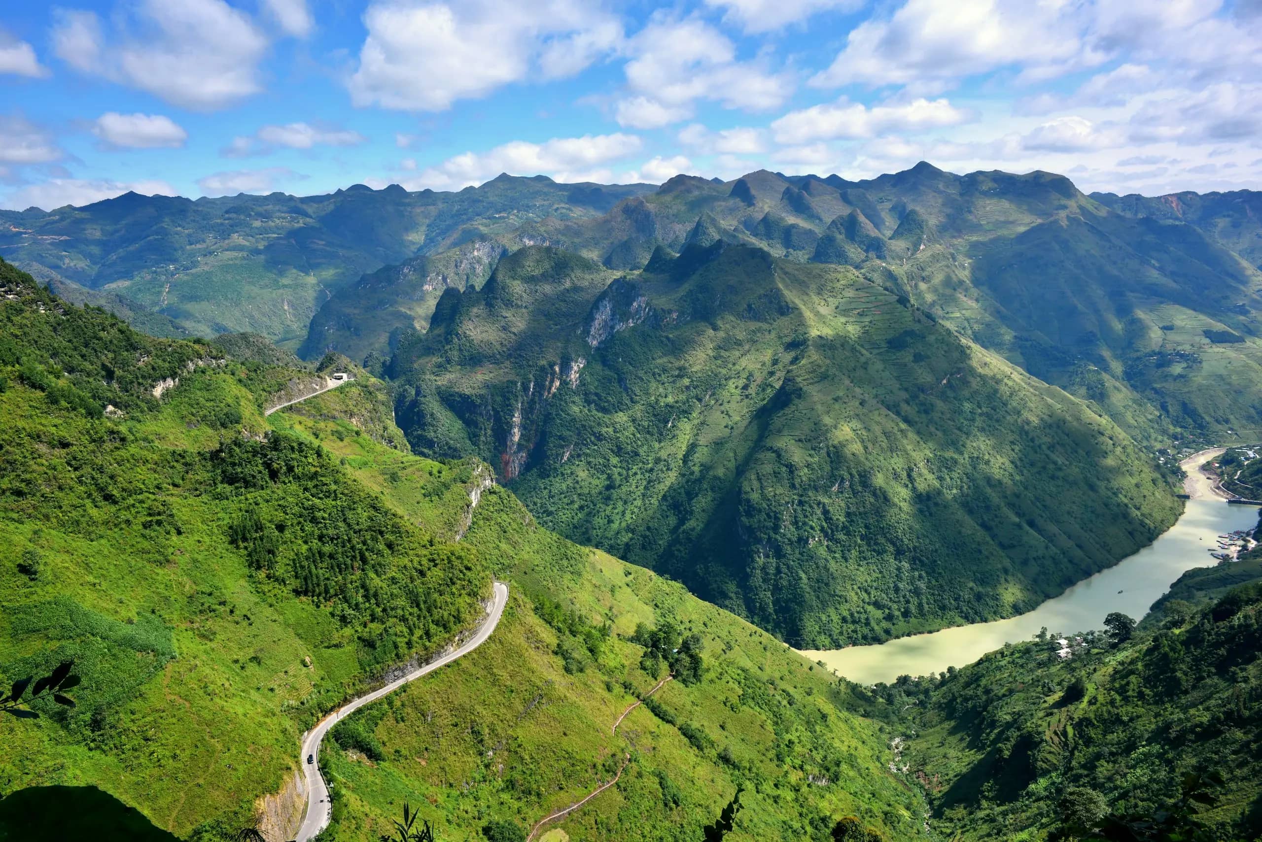 A narrow road winds along green Ma Pi Leng slopes above the jade Nho Que River, karst peaks under a blue sky