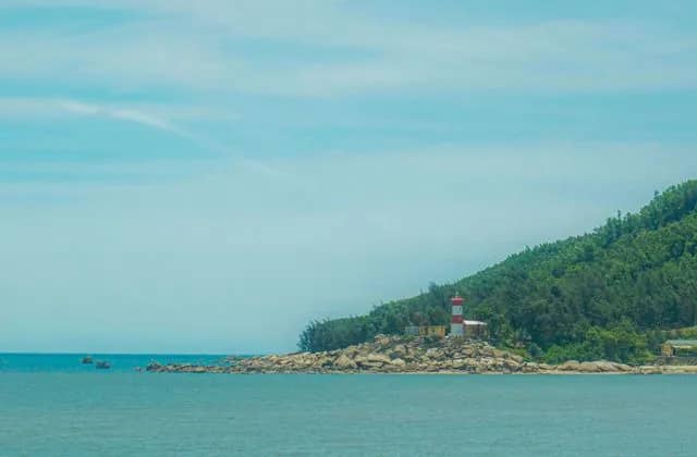A red and white lighthouse on a rocky, forested headland above the calm blue sea of the Ha Tinh coast