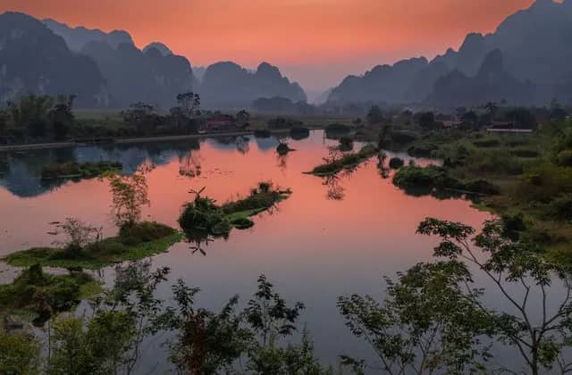 Karst mountains silhouetted against a pink sunset sky above a still, winding lake and green islets in Lang Son