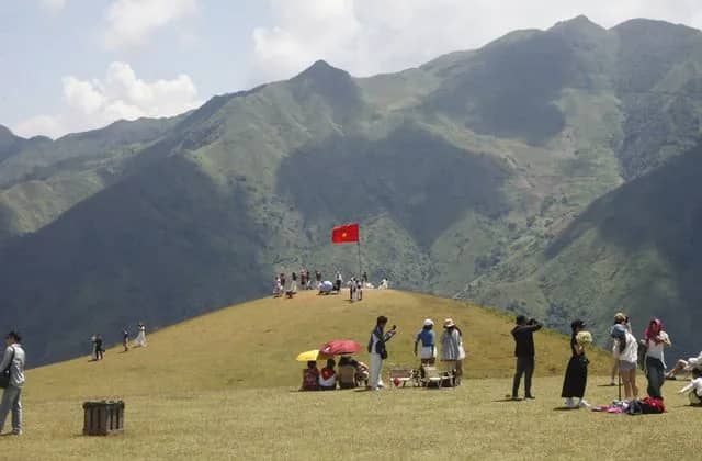 Visitors on a grassy summit at Ta Xua, a flag flying, mountain ridges behind
