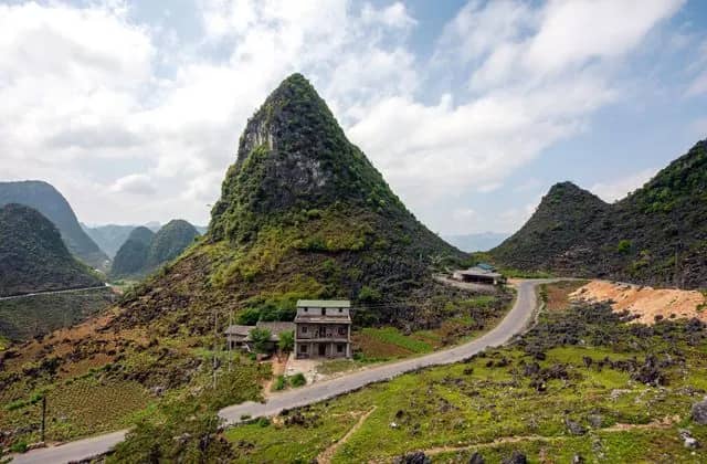 A pointed karst peak rises over a roadside house and a winding road in the mountains of Ha Giang