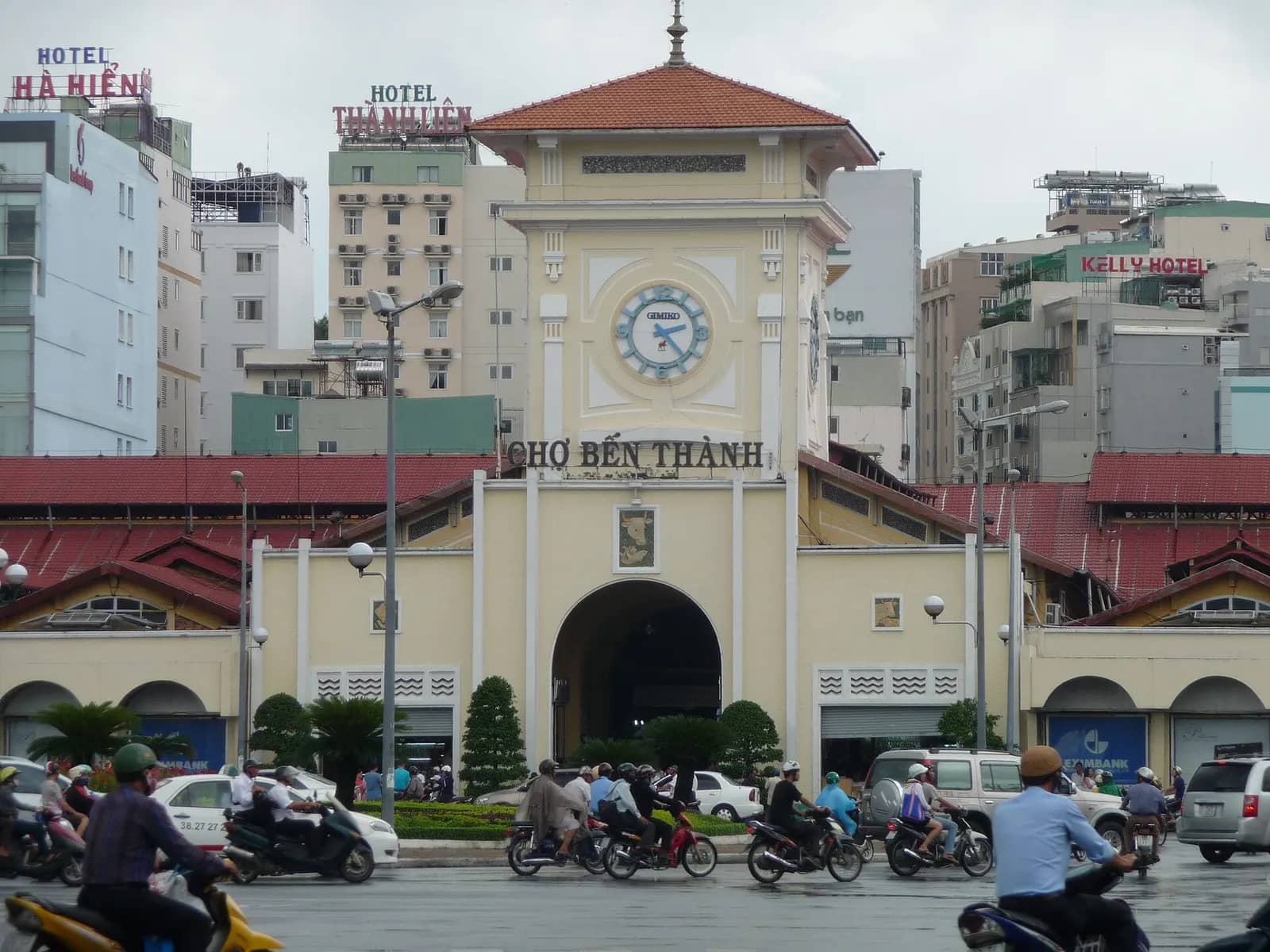 Cream front gate of Ben Thanh Market with its clock tower and Cho Ben Thanh sign, motorbikes passing on the road