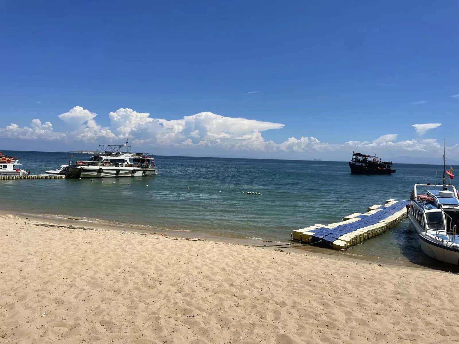 A sandy beach on Cu Lao Cham with two passenger boats moored at a floating pier, blue sea and white clouds behind