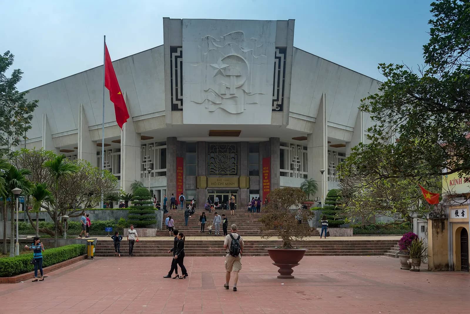 White Ho Chi Minh Museum building with a carved emblem above the entrance, a red flag on the left and visitors on the forecourt