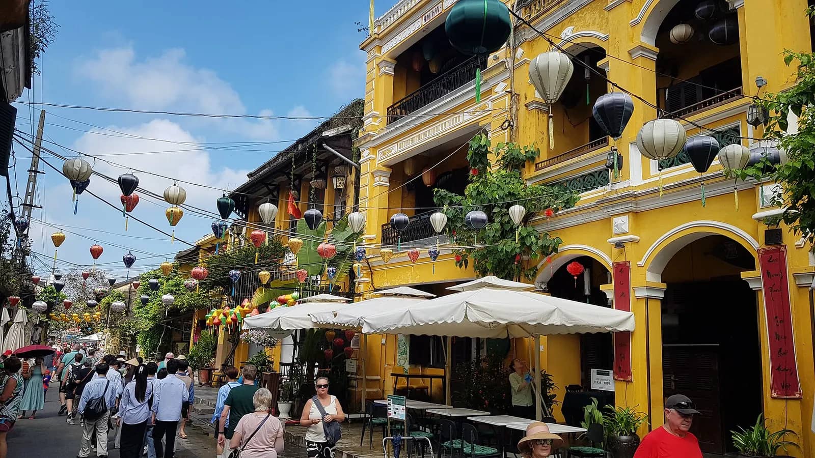 A busy yellow-walled street in Hoi An Ancient Town hung with lanterns, with visitors walking under cafe umbrellas