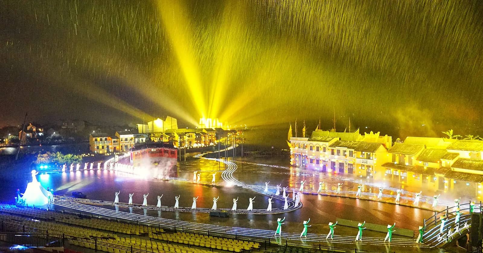 Rows of performers in white ao dai on a lit river stage during heavy rain, with yellow searchlights over a row of old-style shophouses and empty yellow seats in the foreground