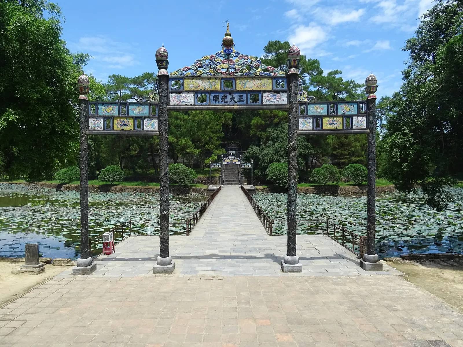 An ornamental gateway on a stone causeway that crosses a lotus pond at Minh Mang Tomb, with trees behind
