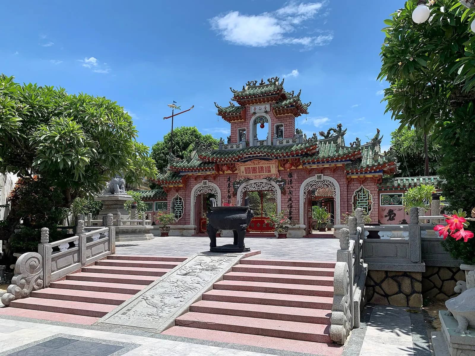 The pink and green triple gate of Phuc Kien Assembly Hall seen across a paved courtyard with stone steps, a bronze incense urn and trees under a blue sky