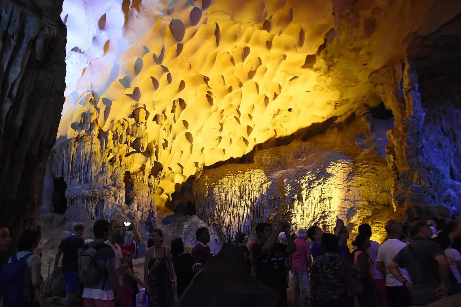 Visitors crowd a floodlit chamber in Sung Sot Cave under a yellow ceiling pitted with scallop-shaped hollows