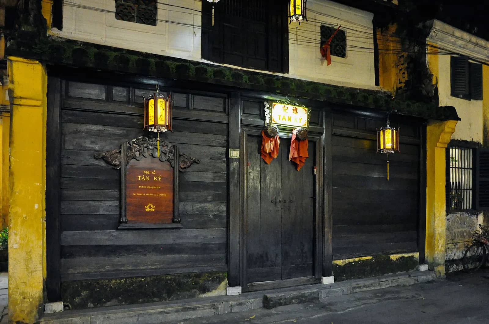 The dark wooden shopfront of Tan Ky Old House at night, with a carved name board, two hanging lanterns and yellow walls on either side