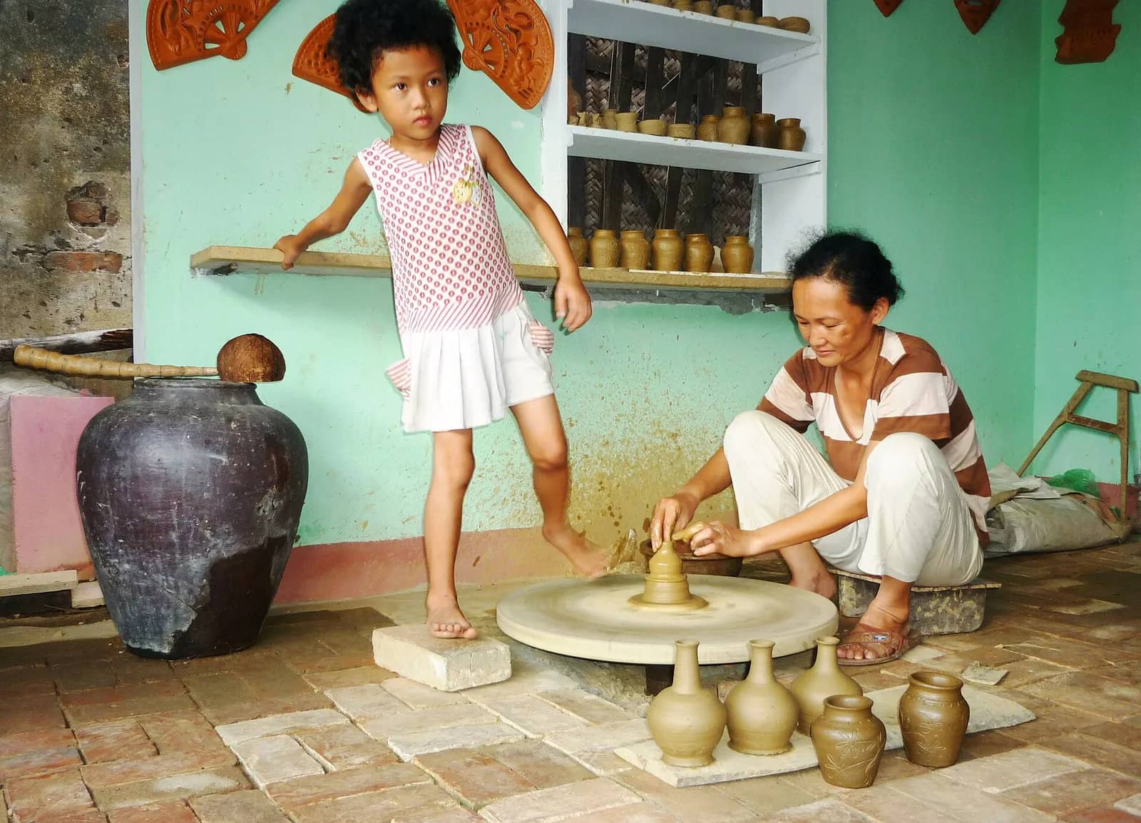 A woman shaping a small pot on a low potter's wheel while a young girl stands on the wheel's edge turning it with her foot, finished pots around them