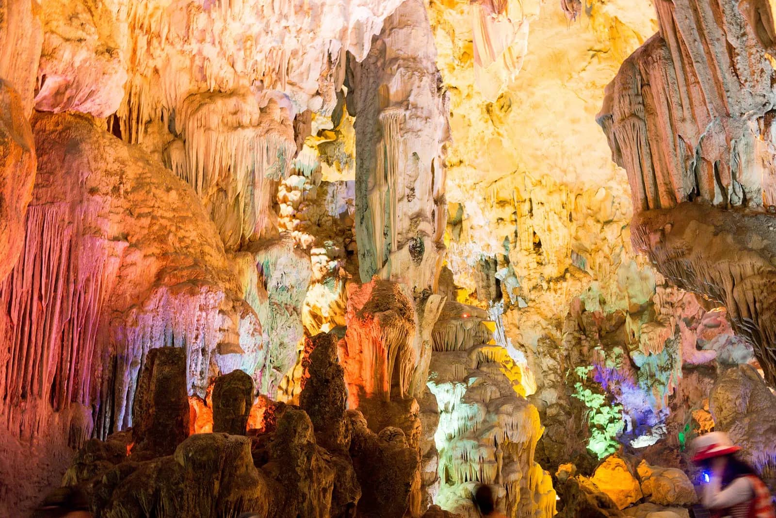 Stalactites and stalagmites in Thien Cung Cave lit in orange, white and violet, with a visitor at the lower right
