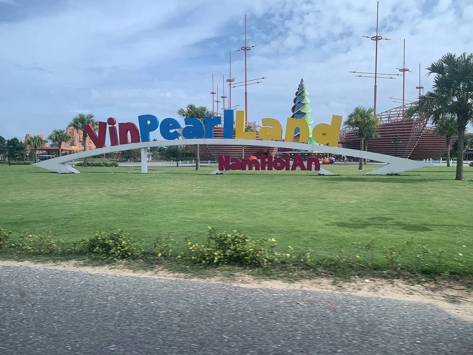 A large colorful Vinpearl Land Nam Hoi An sign on a lawn beside a road, with the masts of a replica sailing ship and palm trees behind it