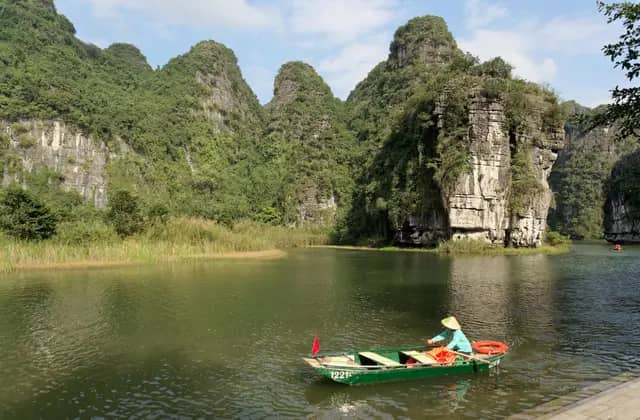 A rower guiding a small boat across a calm lake below limestone peaks in Ninh Binh