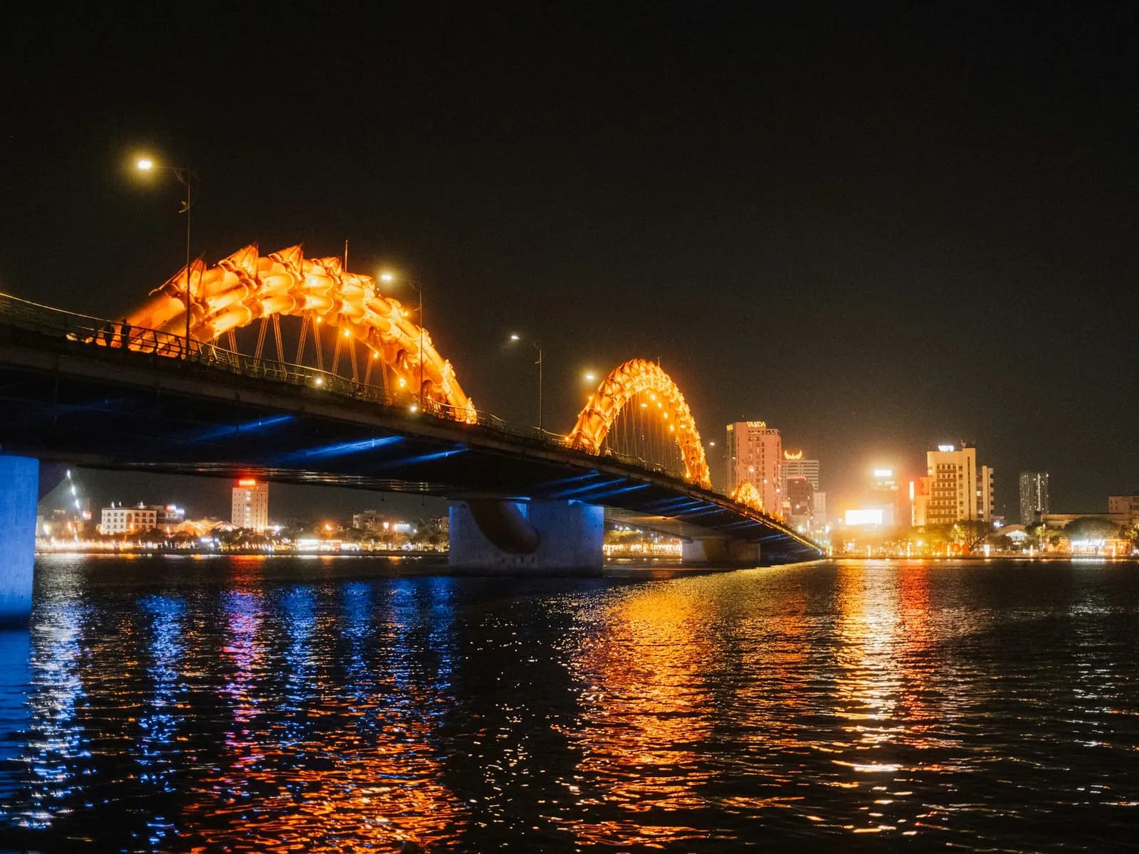 The Dragon Bridge in Da Nang lit orange at night, its arches and city lights reflected in the Han River