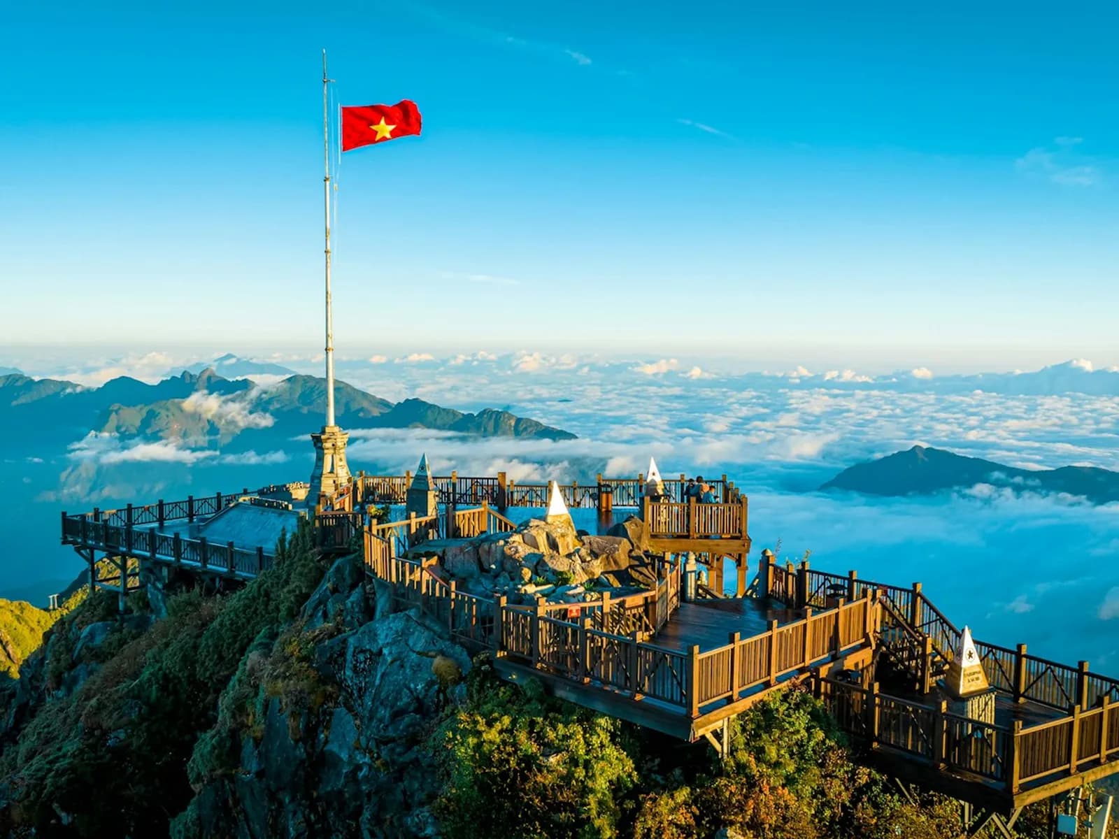 A red Vietnam flag on the Fansipan summit, wooden viewing decks below and a sea of cloud under blue sky
