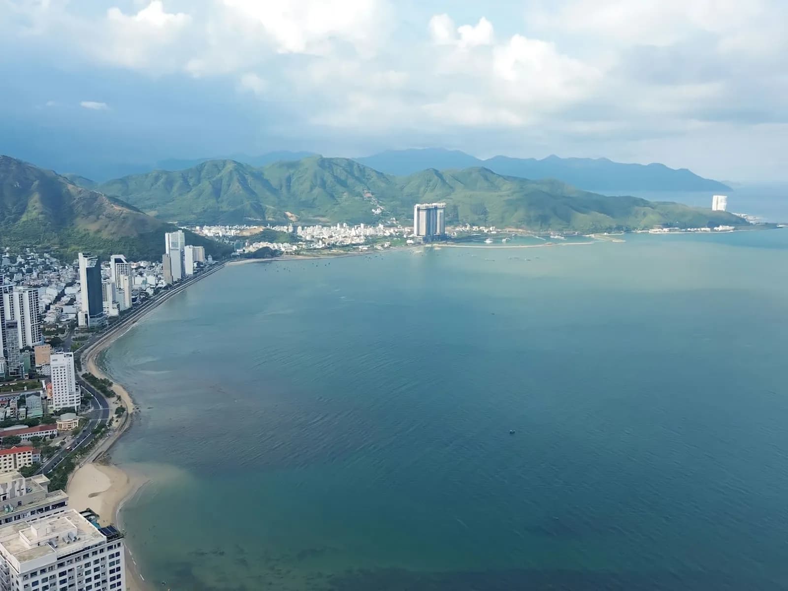 Aerial view of Nha Trang Bay, with hotel towers along the curving seafront and green mountains under white clouds