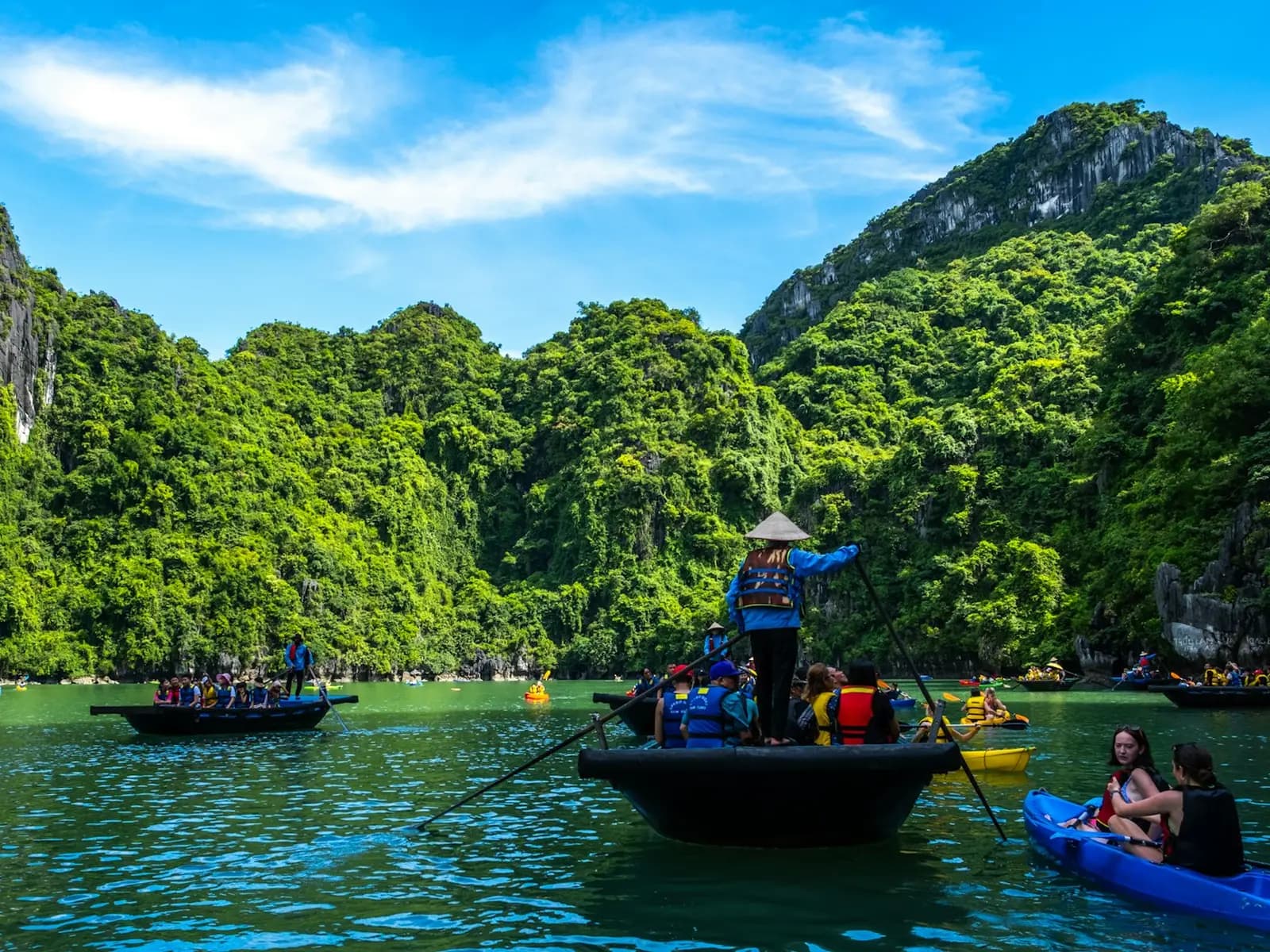 A rower in a conical hat steering a bamboo boat of visitors past kayakers below green karst cliffs in Ha Long Bay