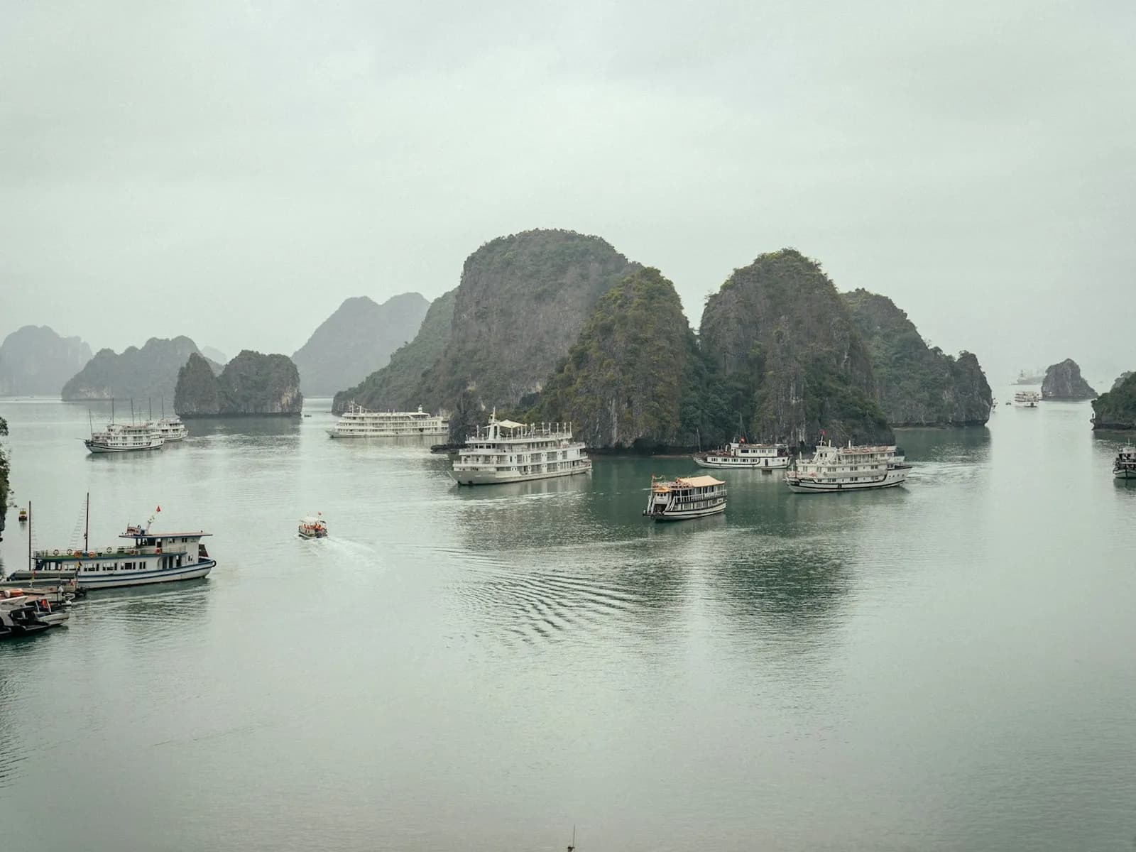 Cruise boats anchored on grey-green water among limestone islands in Ha Long Bay on a misty, overcast day