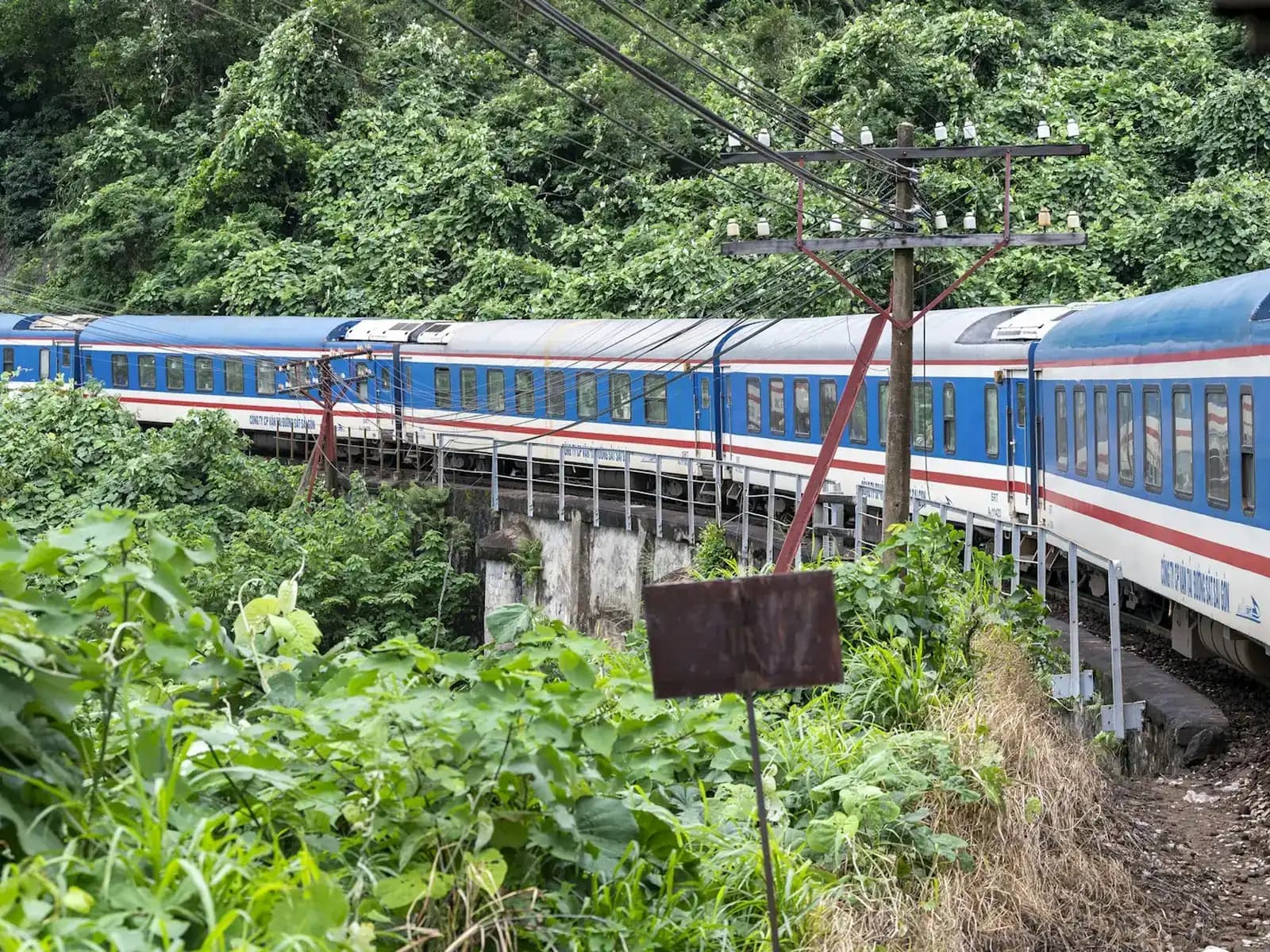 A blue and white Vietnam Railways passenger train on the North-South line curving through dense green forest
