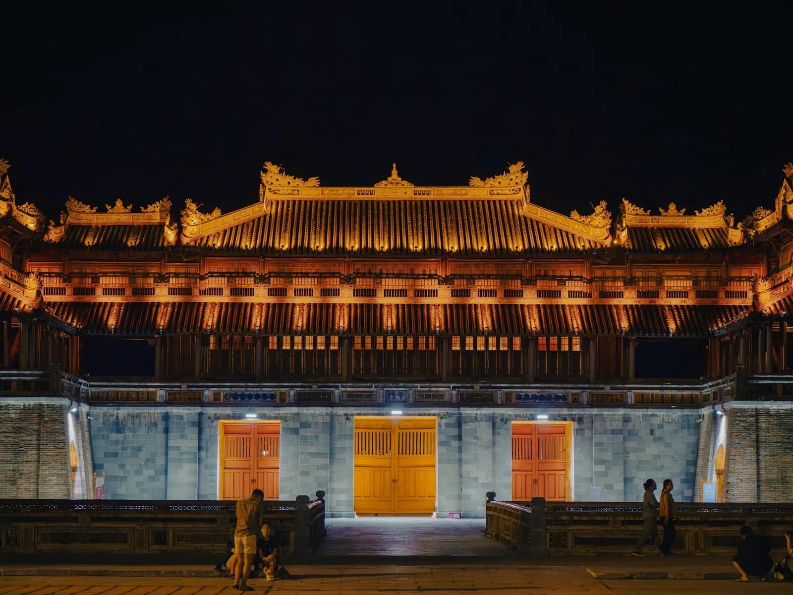 Ngo Mon gate of Hue's Imperial City lit gold at night, with its three doors open and visitors in front