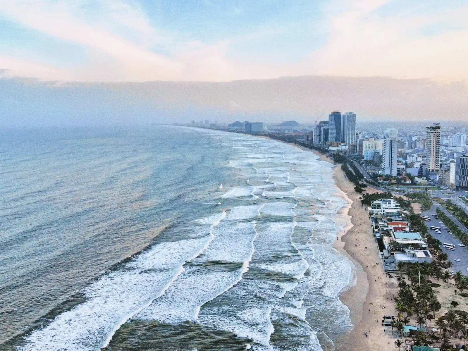 Aerial view of the long Da Nang beachfront at dusk, white surf rolling in beside hotel towers and palm-lined streets