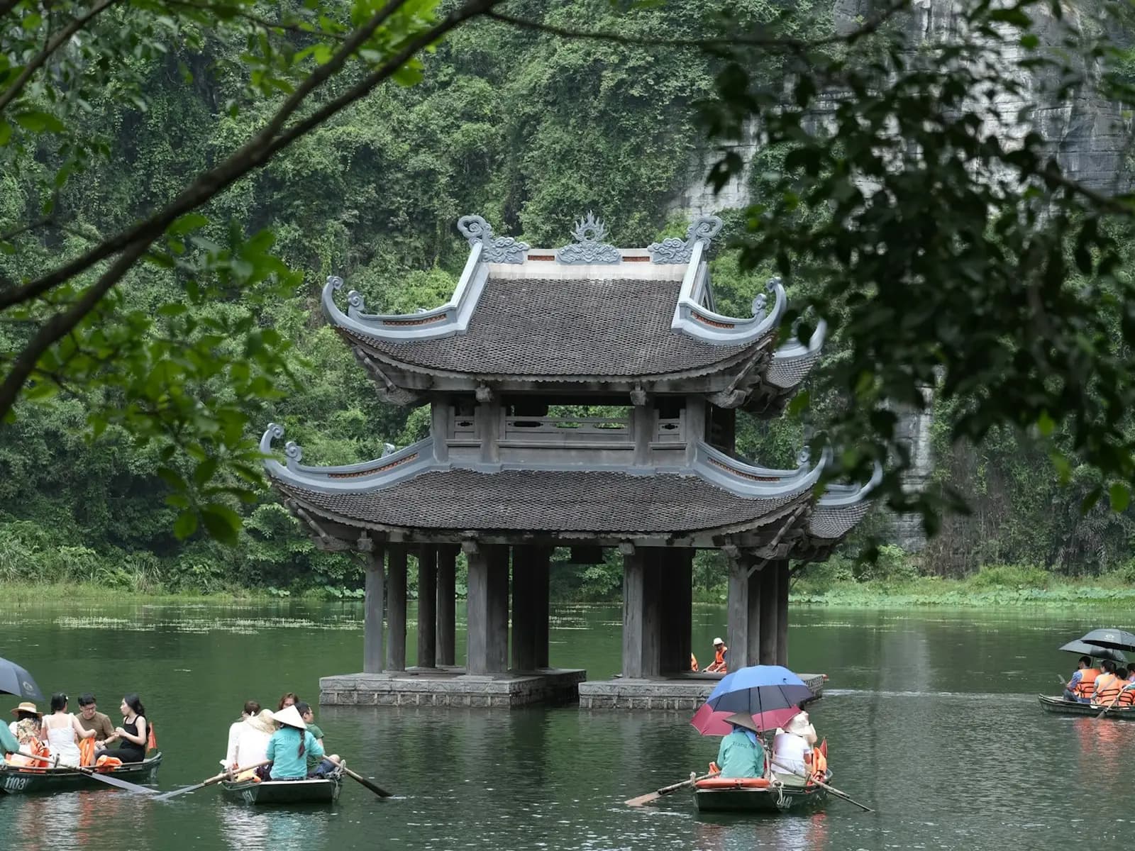 Visitors in rowing boats, some under umbrellas, gliding past a two-tiered pagoda pavilion on the water at Trang An