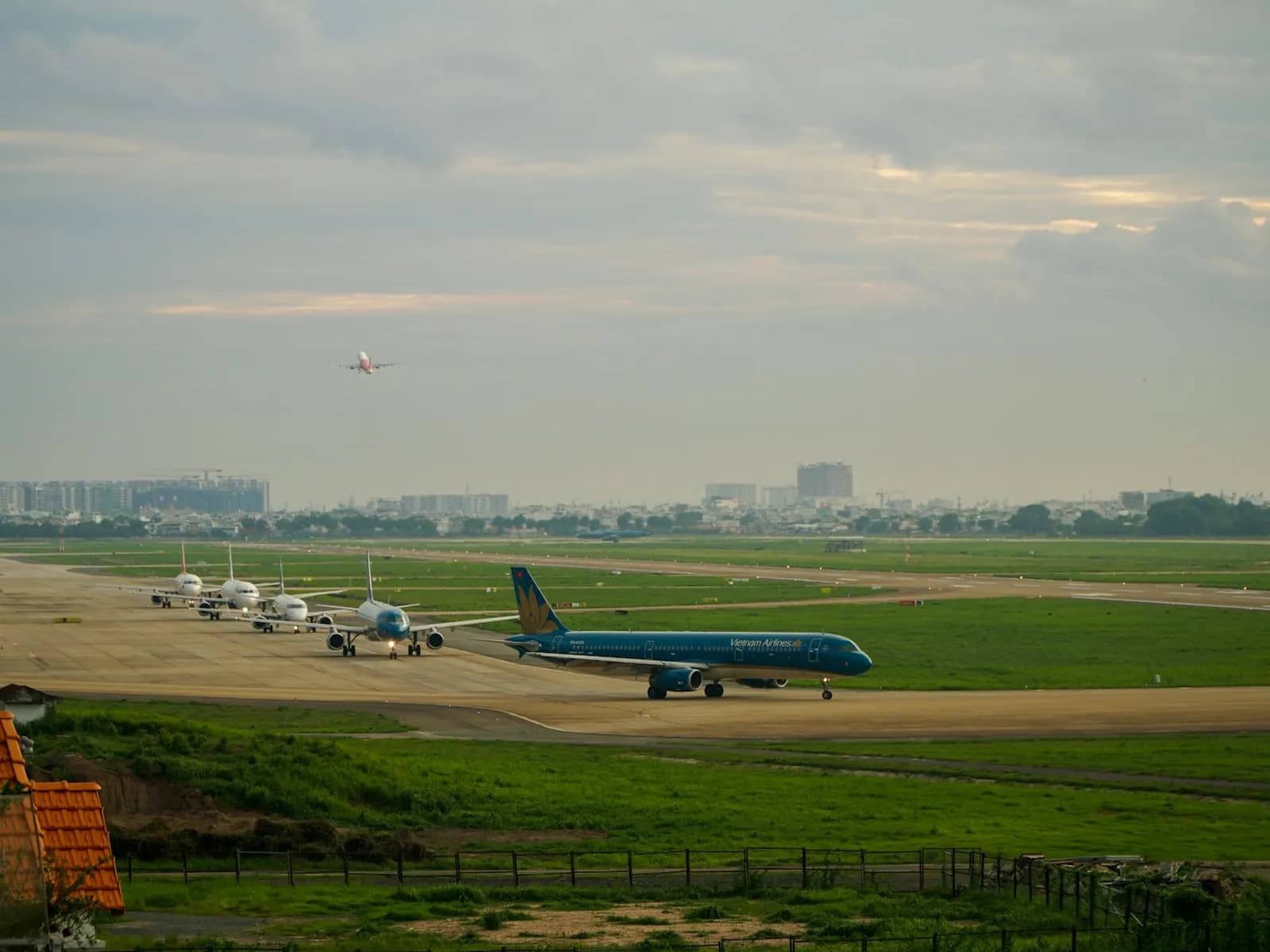 A Vietnam Airlines jet taxiing past a row of parked airliners at Tan Son Nhat airport, with another plane taking off