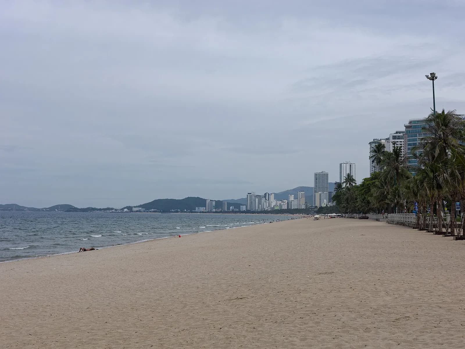 A long, almost empty sand beach in Nha Trang under a grey overcast sky, hotel towers and palms behind