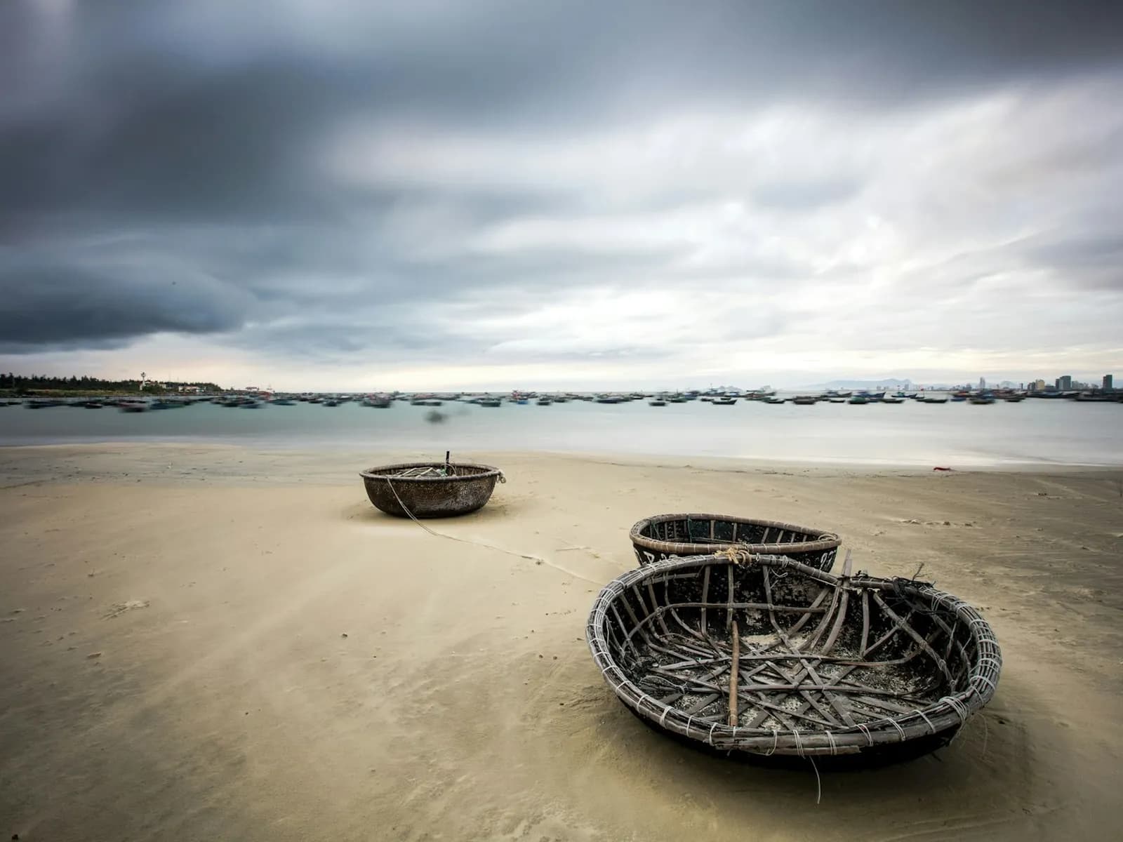 Round bamboo coracle boats resting on a wide sandy beach in Vietnam under dark, heavy storm clouds