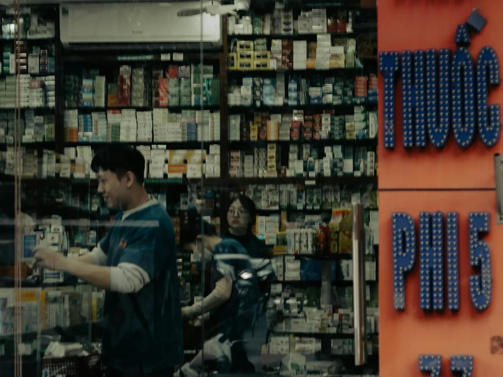 A pharmacist serving customers in a crowded Vietnamese pharmacy, shelves of medicine boxes behind a blue Thuoc sign