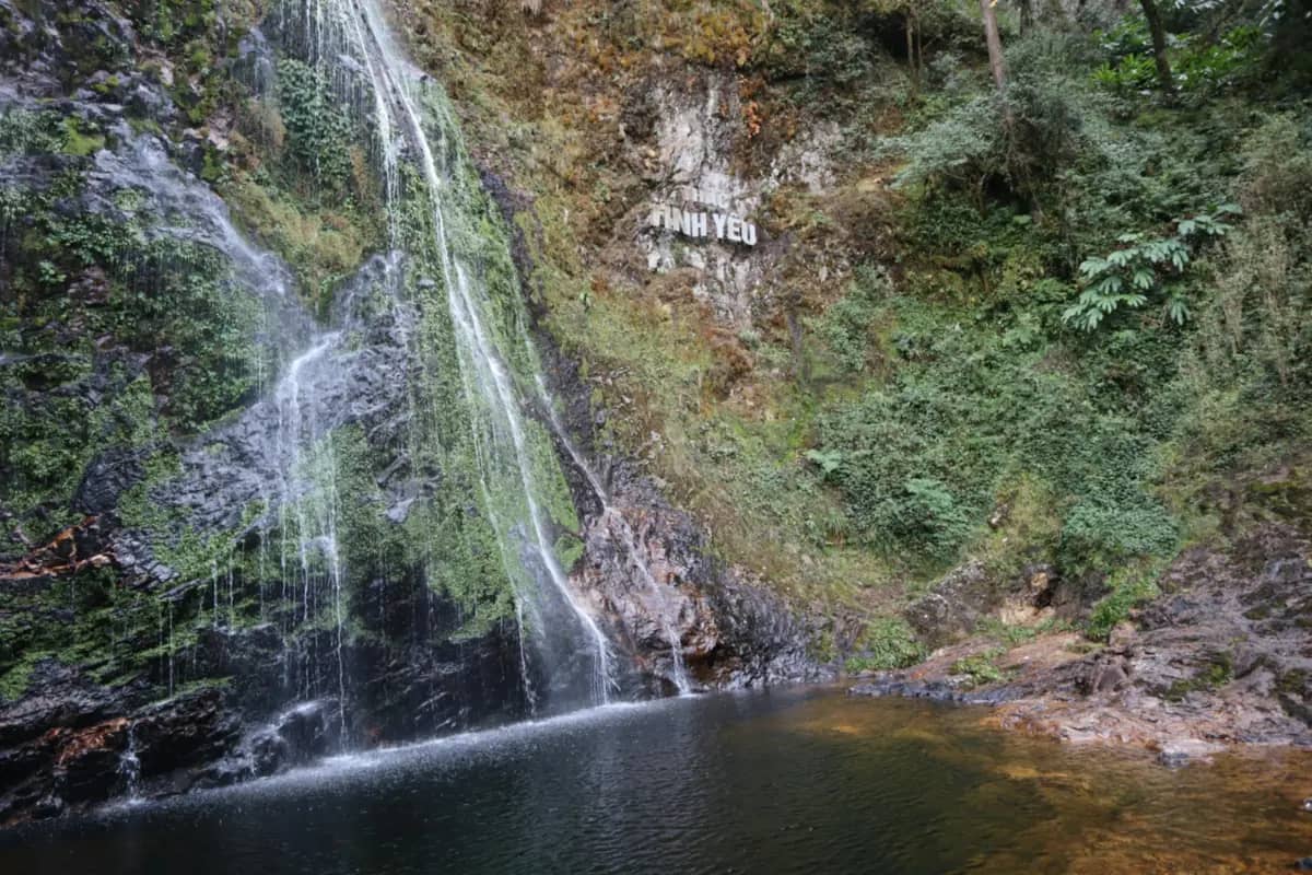 Love Waterfall spilling down a mossy cliff into a dark pool, its name, Tinh Yeu, painted on the rock beside it