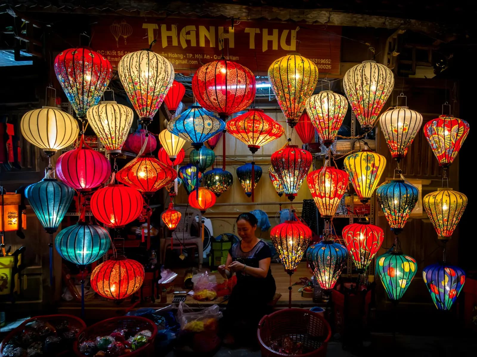 Dozens of glowing silk lanterns in red, yellow and blue hanging over a lantern seller at her stall in Hoi An at night