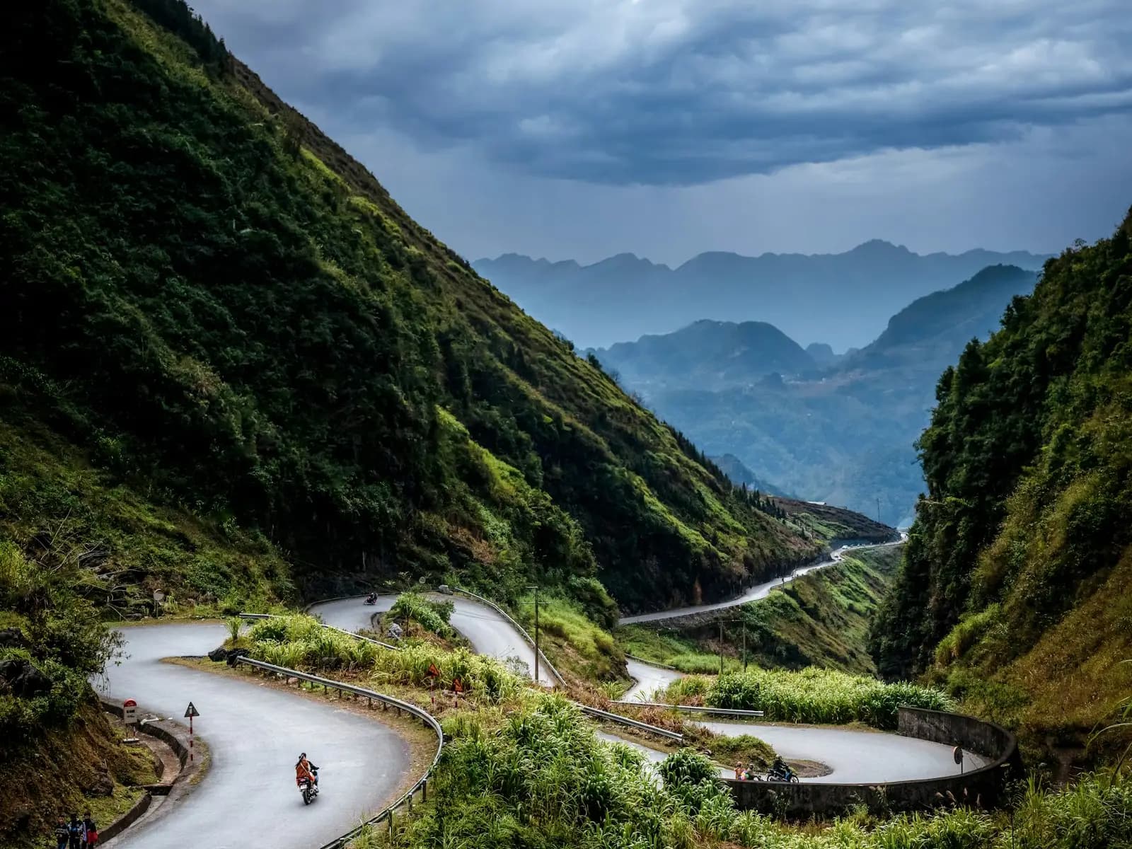 Hairpin road winding down a steep green valley in Ha Giang, with a motorbike rider on the bends below storm clouds