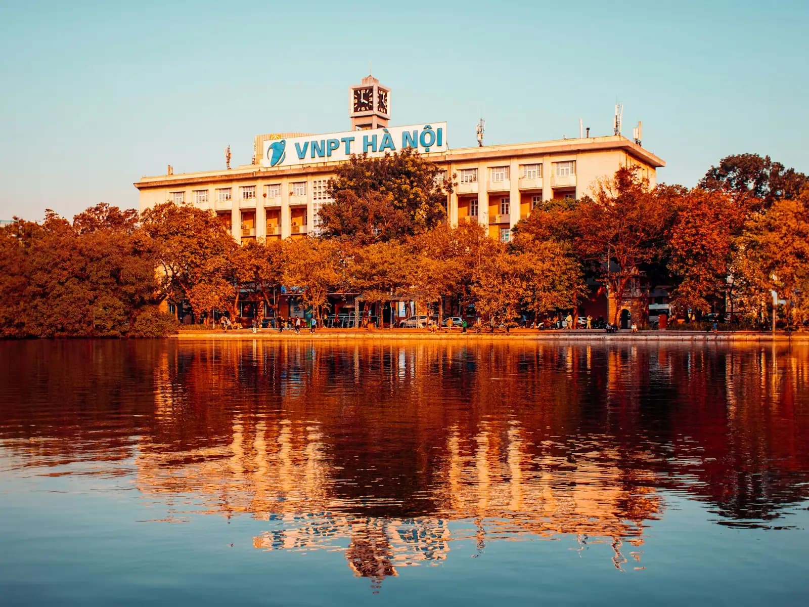 The VNPT Ha Noi building and orange trees reflected in calm Hoan Kiem Lake in warm evening light