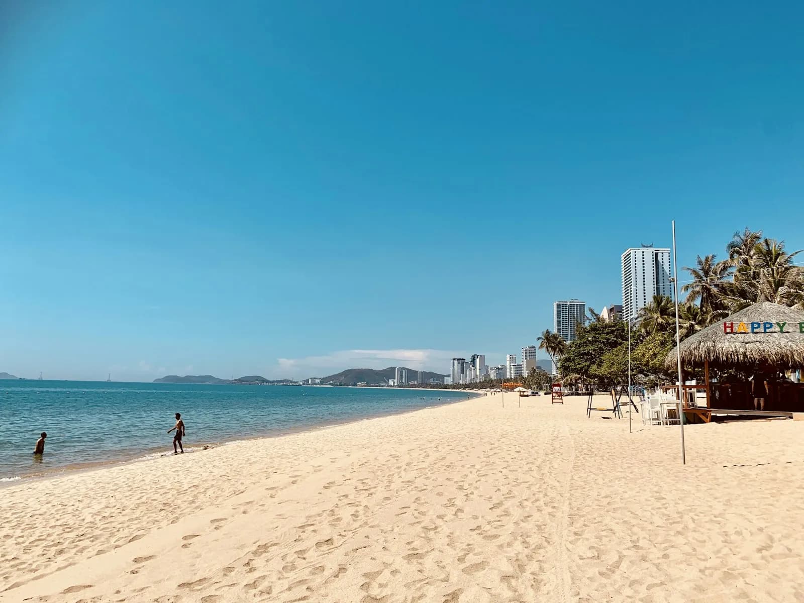 A wide pale sandy beach in Nha Trang under a clear blue sky, with swimmers, palms, a thatched bar and hotel towers