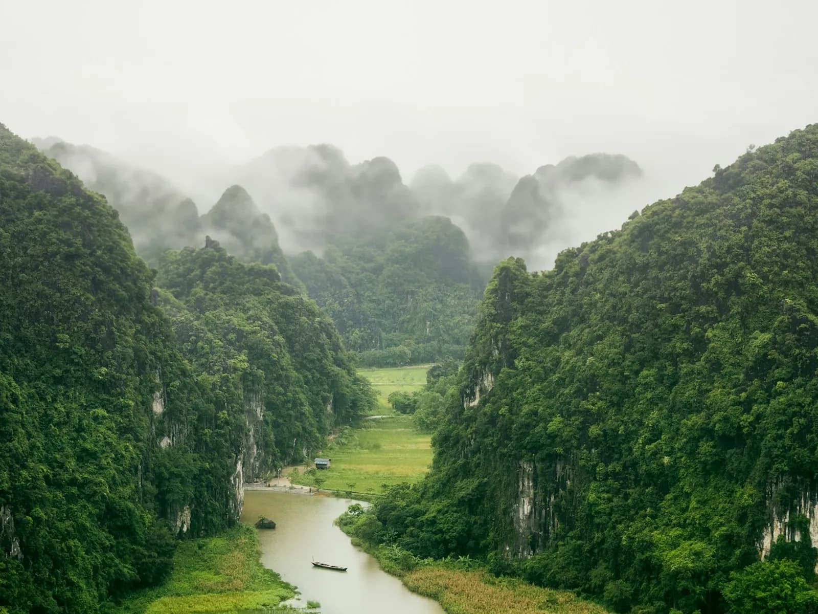 Low mist over forested limestone karsts in Ninh Binh, a small boat on the river winding through green fields