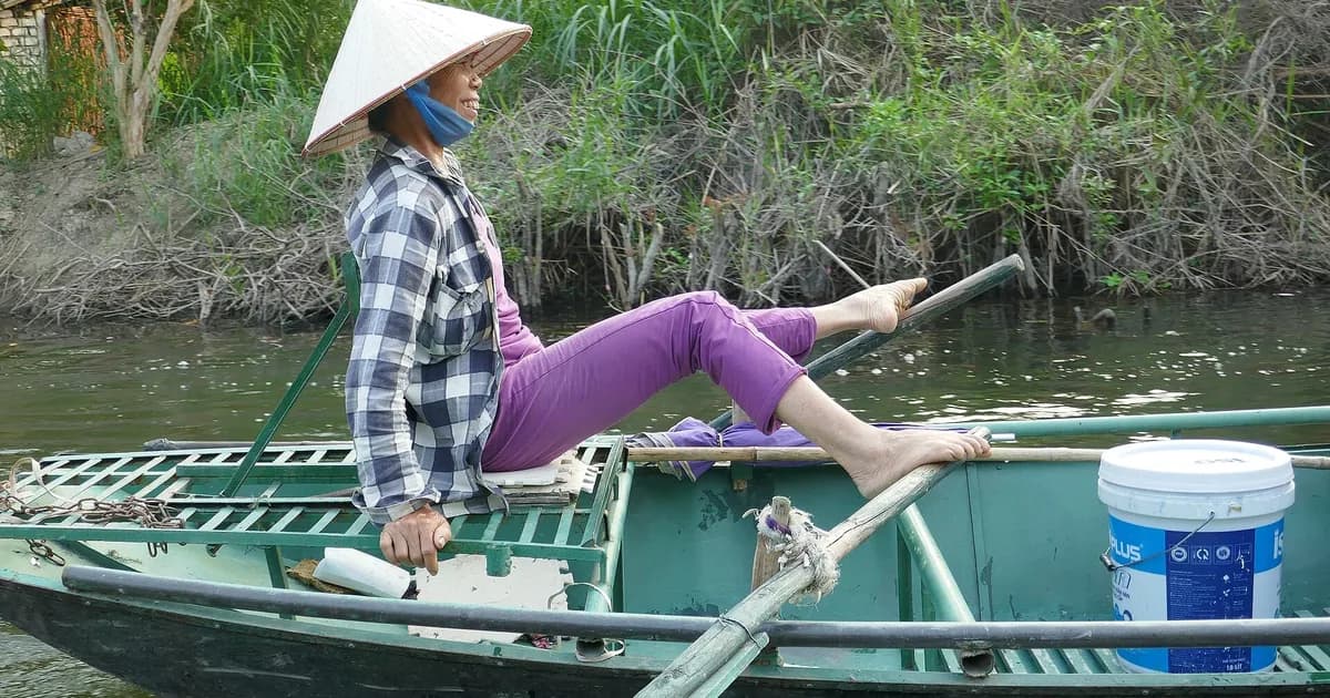 A woman in a conical hat rowing a small green boat with her feet along a riverbank at Tam Coc, Ninh Binh