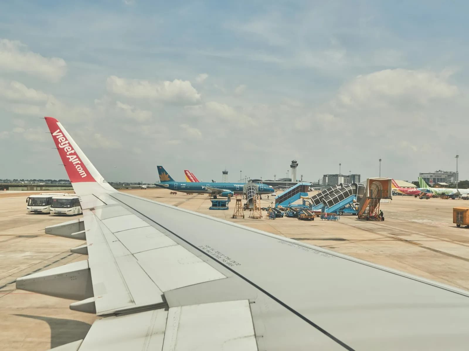 View over a VietJet wing onto the Tan Son Nhat apron, with Vietnam Airlines jets, boarding stairs and the control tower