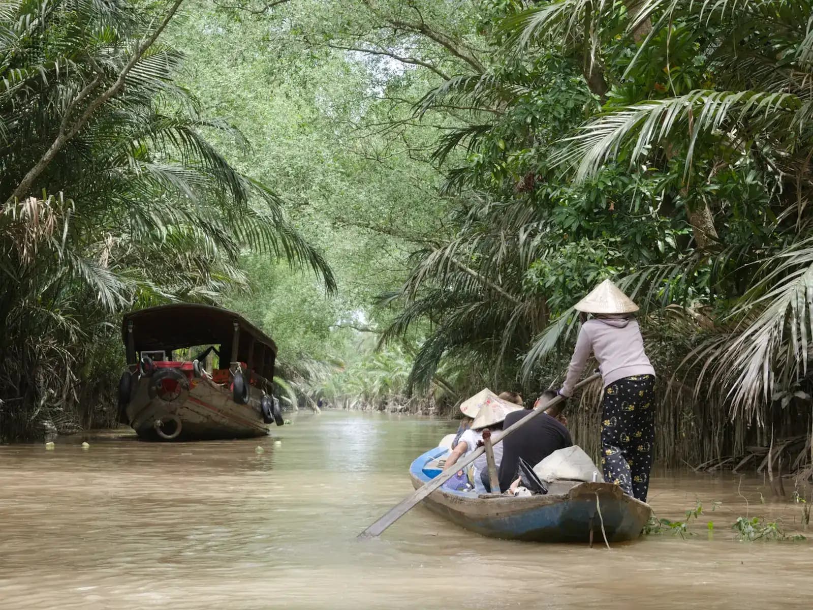 A woman in a conical hat rowing visitors along a brown Mekong Delta canal lined with water palms