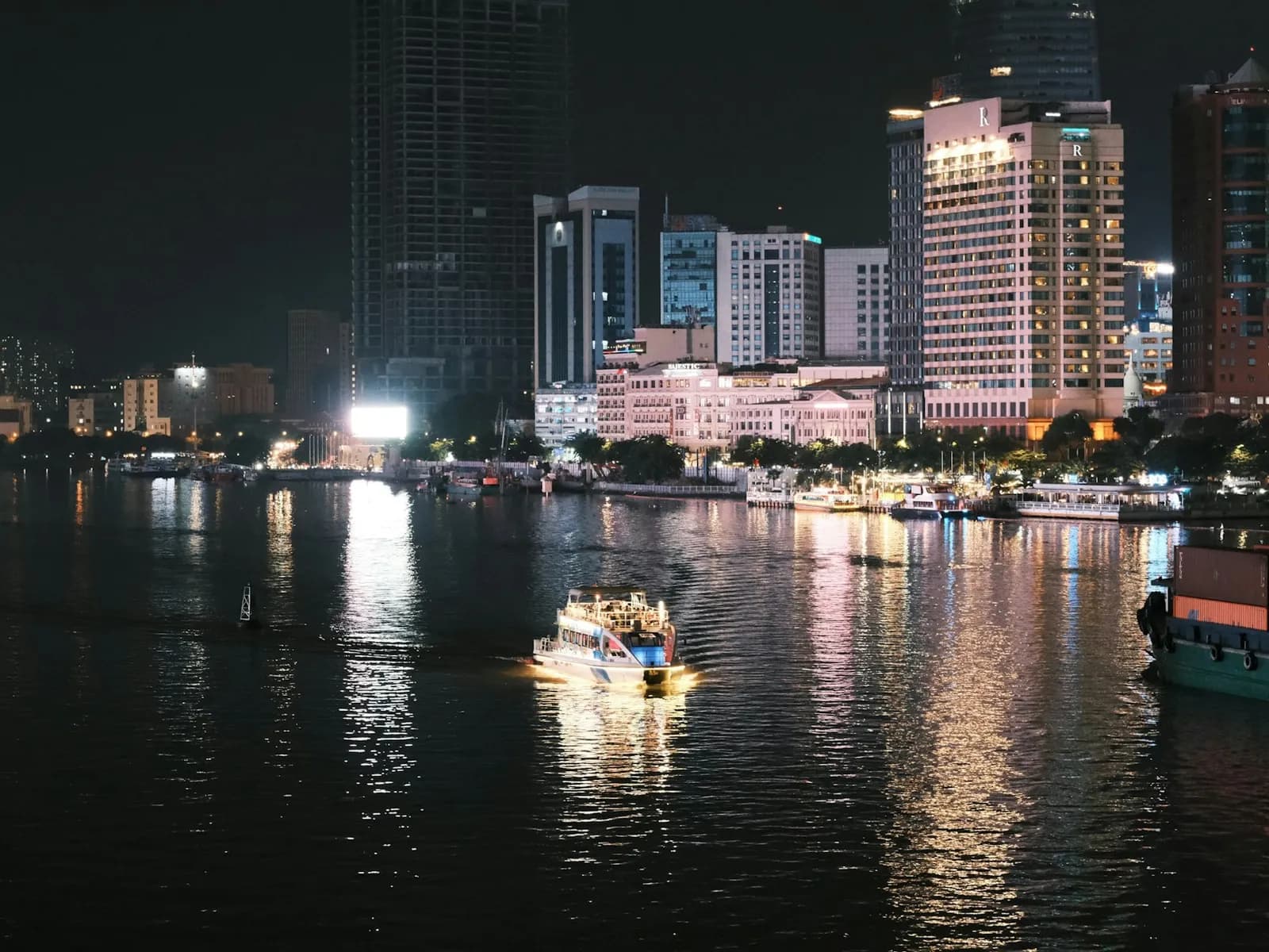 A lit sightseeing boat on the Saigon River at night, with hotel towers of downtown Ho Chi Minh City on the bank