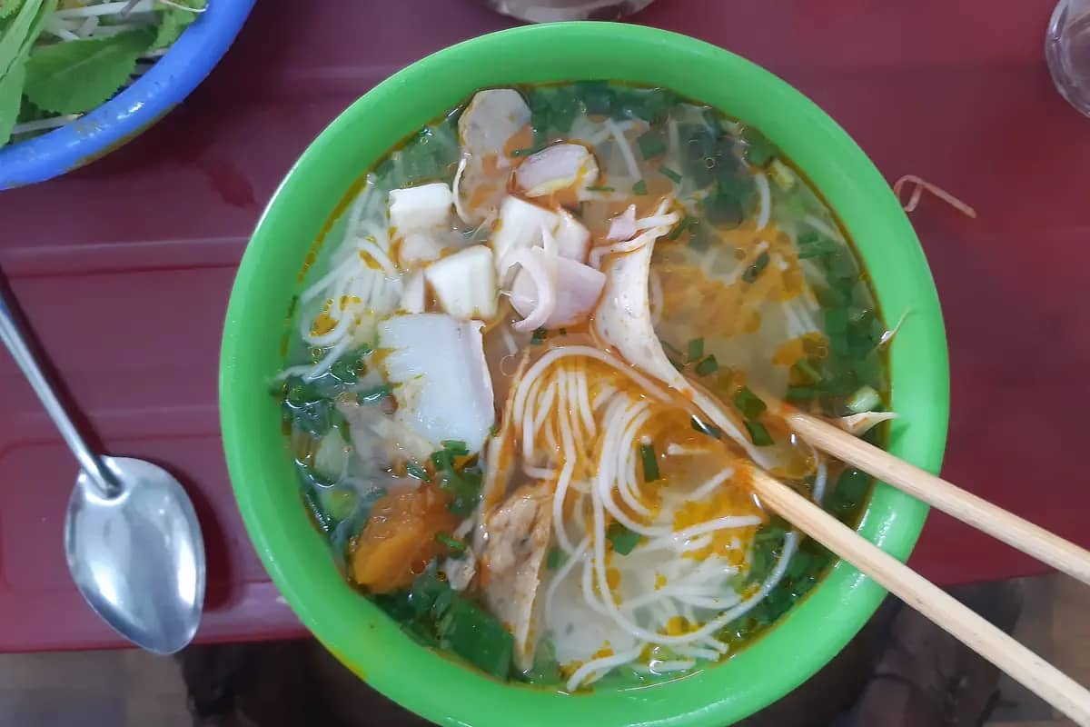 A bowl of bun cha ca: rice noodles with sliced fishcake in a red broth, chopsticks resting on the rim