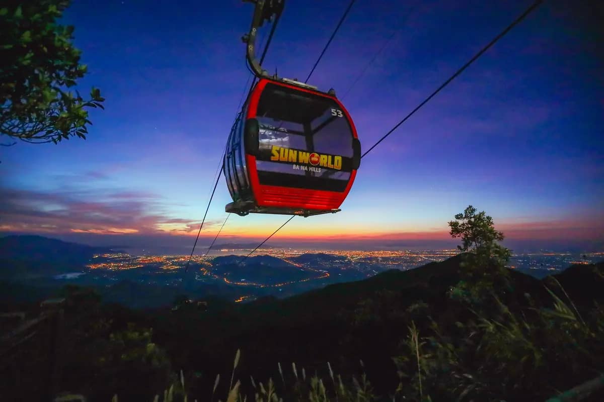 A red Sun World cable car cabin above the forested slopes of Ba Na Hills at dusk, city lights below