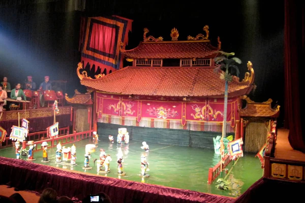 Wooden water puppets gliding across the pool in front of the red pagoda stage at Thang Long theatre, musicians at the left