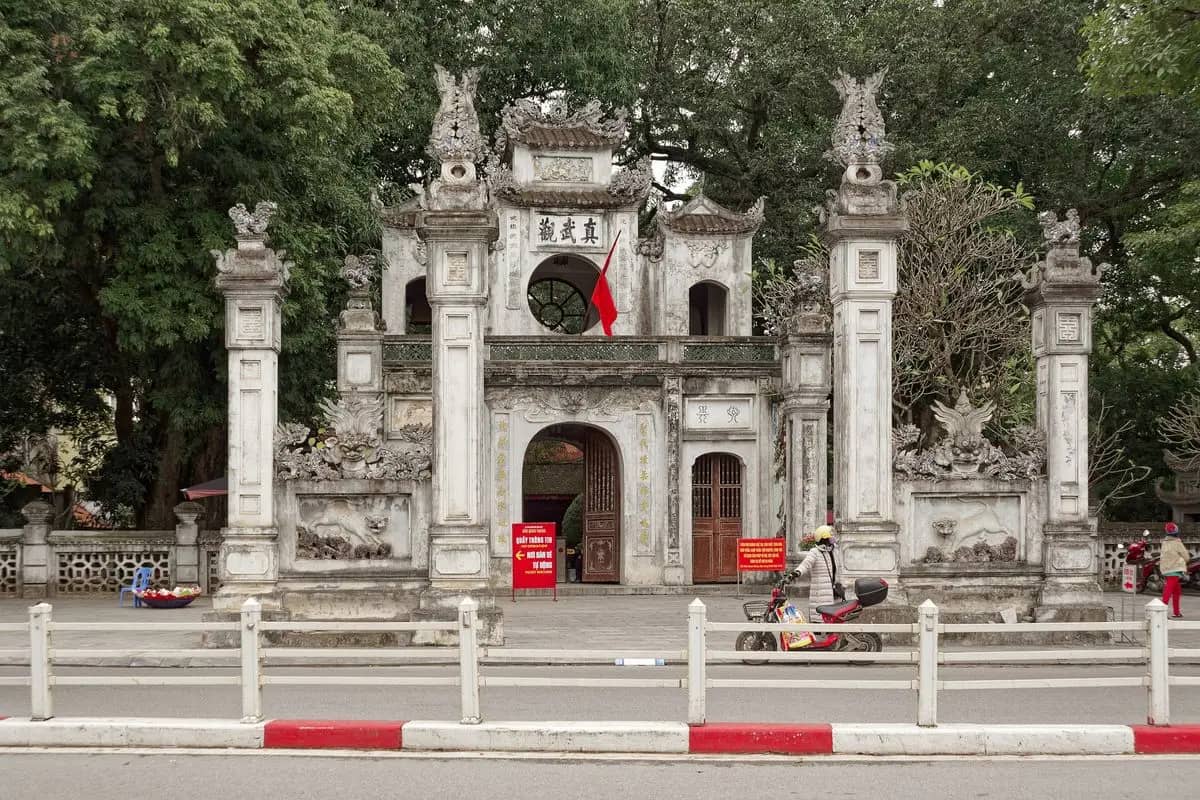 The white stone gate of Quan Thanh Temple under old trees, a red flag in its round window