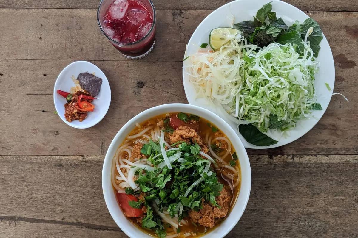 A bowl of bun rieu, crab and tomato noodle soup topped with herbs, beside a plate of shredded greens