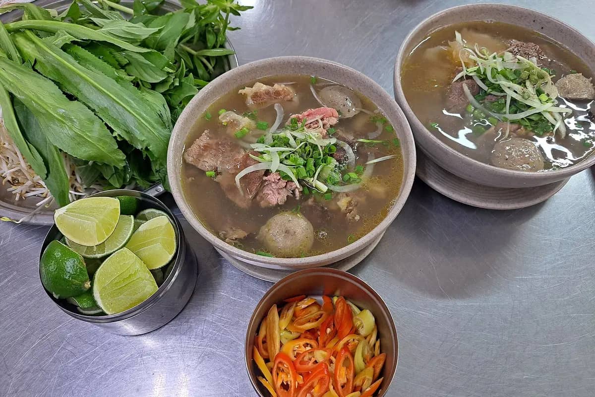 Two bowls of pho with beef and beef balls, herbs, bean sprouts, lime and chilli on the side